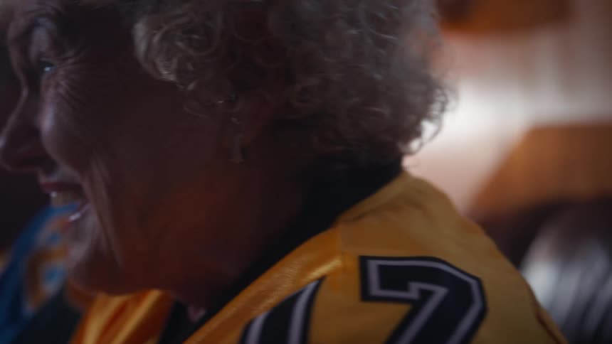 An elderly couple sits on a couch in the living room at midday wearing jerseys and watching a sports event with excitement
