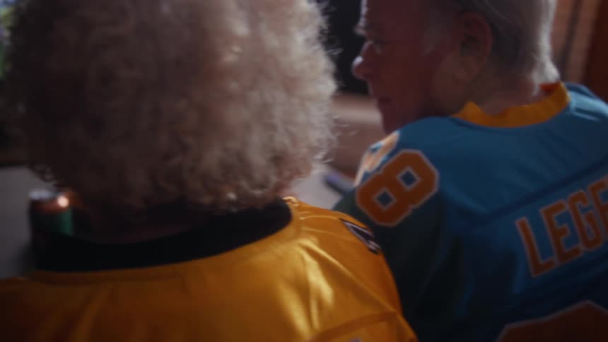 The video shows the backs of two elderly people sitting close together and watching a game on tv with the mans blue jersey featuring legend and the number 88 the foreground is dominated by the curly hair of the woman in the yellow jersey as they both