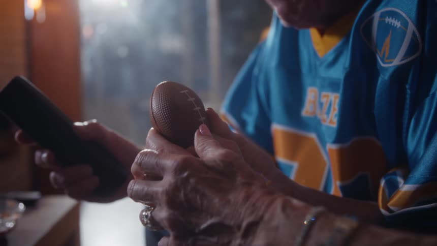 A closeup shot focuses on the aged hands of two people one of whom is wearing a bright blue and yellow sports jersey as one hold a miniature football while the other person holds a black remote control