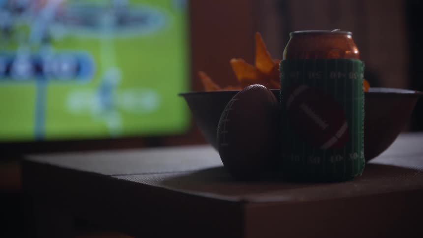 Beverage can and a bowl of chips sit on a table in the living room while sports play on a television