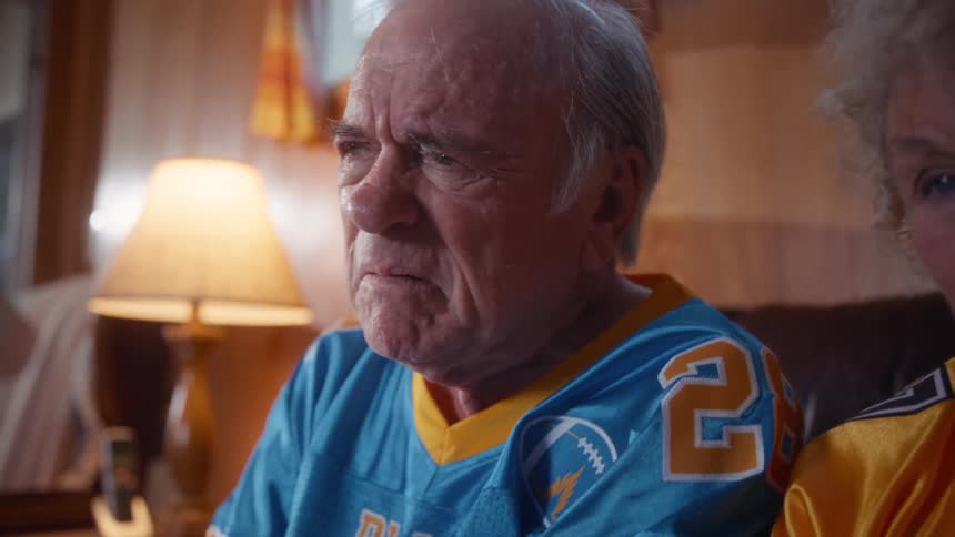 The shot quickly moves from a closeup of an elderly woman with her eyes closed to focus on an elderly man wearing a blue football jersey with a distressed expression while looking intently offcamera