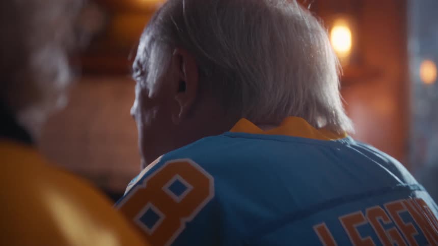 A shot focuses on the backs of two older individuals wearing sports jerseys with one wearing a blue jersey labeled legend 88 and the other wearing a yellow jersey labeled rookie they appear to be engaged in conversation in a dim indoor setting pos