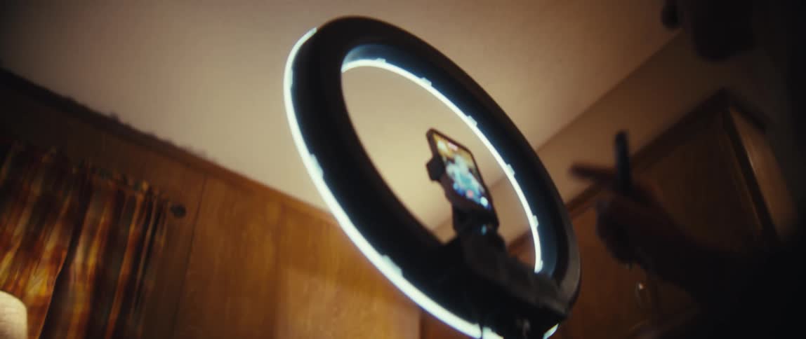 The focus shifts from an elderly woman holding a trading chart to the smartphone recording her on a ring light