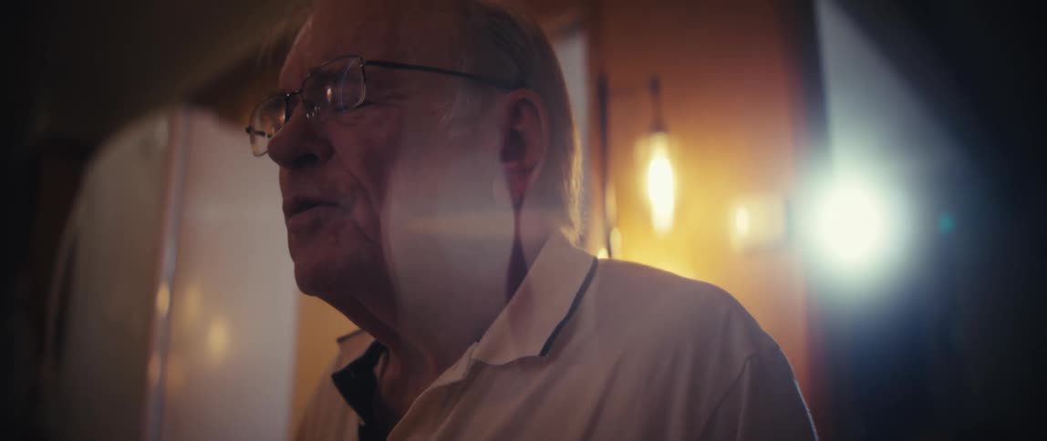 A closeup shot captures an elderly man wearing glasses and a white polo shirt as he speaks with emotion to someone offcamera in a dimly lit room