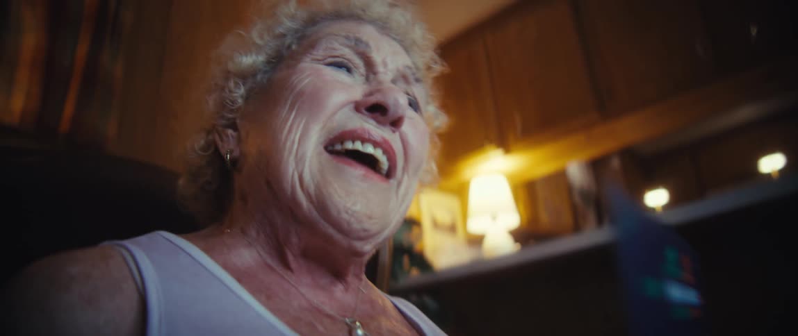 An elderly woman with curly lightcolored hair wearing a white top and a pearl necklace holds up a chart detailing candlestick trading patterns in a dimly lit woodpaneled indoor setting
