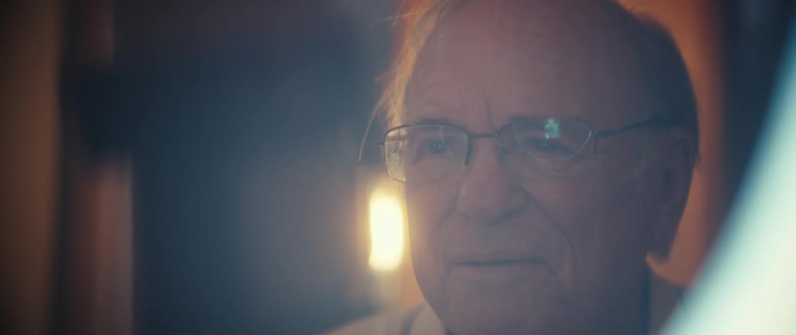 A closeup shot focuses on the face of an older man wearing glasses whose expression is calm and engaged suggesting he is listening or conversing the lighting is dim and warm possibly reflected off a screen or another light source creating a soft ha