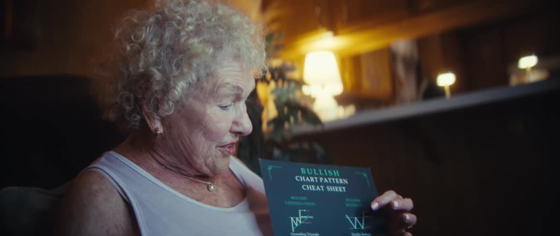 The video opens with a closeup of a smartphone attached to a ring light showing a woman on its screen and then cuts to the woman herselfthe elderly woman with curly hair and a pearl necklace smiles excitedly as she holds up a bullish chart patter