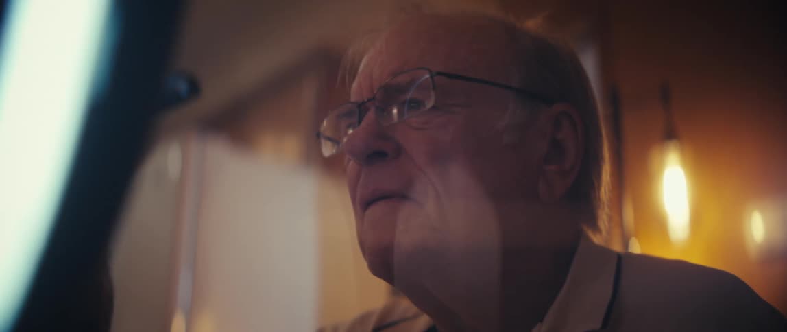 An elderly man wearing eyeglasses typing on a smartphone mounted on a stand with the bright circular ring light reflecting in his glasses