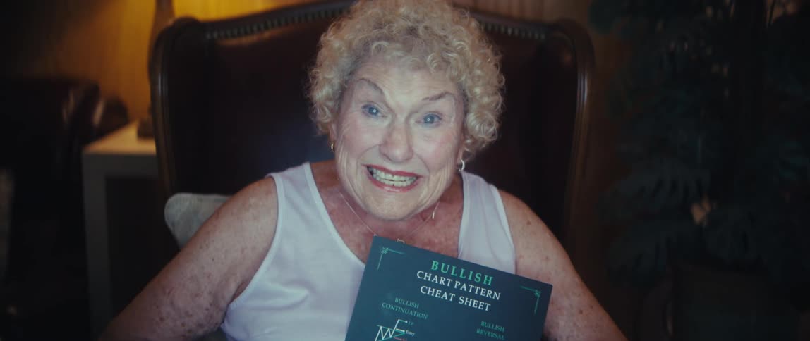 A close view of a cheerful older woman with curly blonde hair and a white tank top sitting in an armchair holding a black bullish chart pattern cheat sheet and smiling brightly