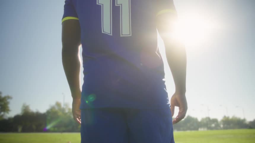 A soccer player wearing a blue jersey marked captain stands on a sunlit field with a ball at his feet