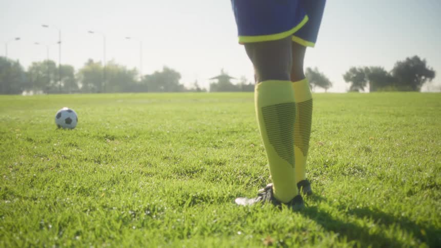 A lowangle shot focuses on the lower legs of a soccer player wearing blue shorts and bright yellow and black socks standing still on a green grass field with a soccer ball resting a short distance away under bright sunlight