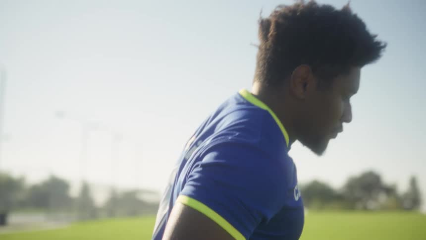 A male soccer player in a blue and yellow jersey and yellow socks dribbles a black and white ball on a green grass field on a bright sunny day