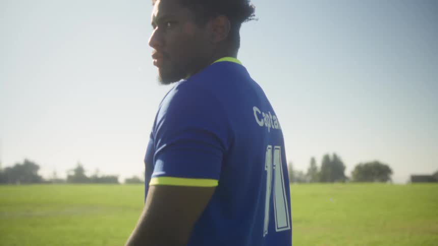 A male soccer player wearing a blue and yellow uniform standing on a green grass field on a sunny day