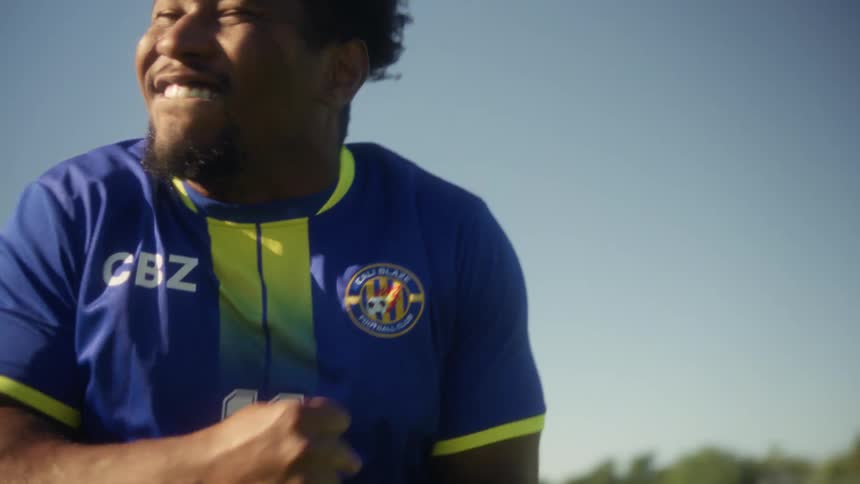 A soccer player in a blue and yellow jersey kicks a ball on a green field raising his fists to celebrate while shouting under a clear blue sky