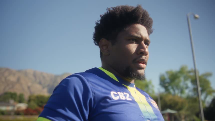 The video shows a closeup shot of a black man with a full afro and a goatee wearing a blue and yellow soccer jersey with cbz printed on it he is standing outside on a sunny day with a mountain range and trees in the background appearing focused and