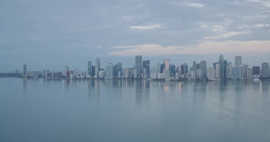 An elevated view of a large modern city skyline reflecting on the flat calm water below the horizon under a pale gray cloudy sky