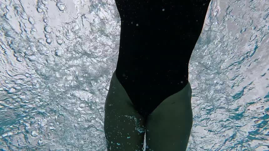 An underwater view shows a woman in a black swimsuit and goggles swimming freestyle under a string of dark flags against a bright cloudy sky