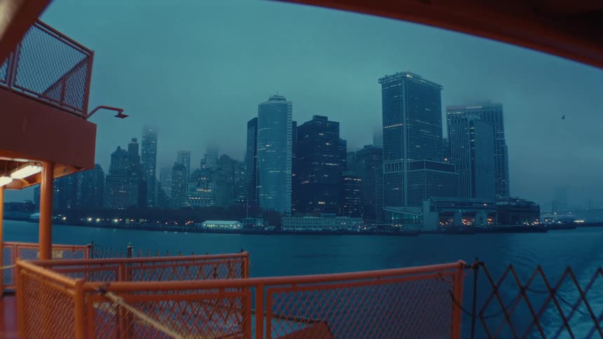 A wide shot of the new york city skyline with dark skyscrapers seen from a ferry deck with orange railings on a cloudy or stormy dark blue evening