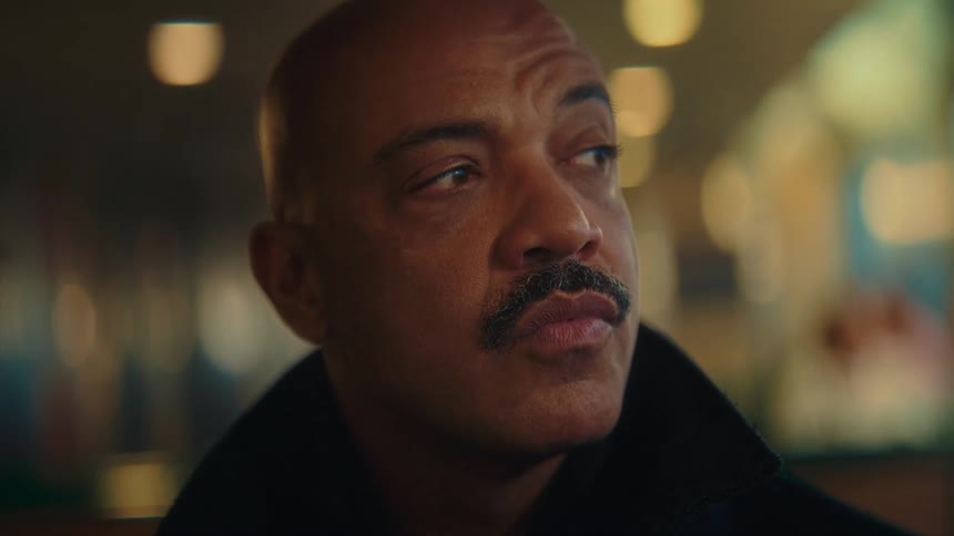 Face of a black man with a mustache wearing a black coat indoors