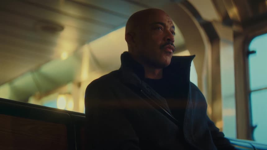 A middleaged man with a shaved head and a mustache is sitting on a wooden bench inside a transit vehicle perhaps a tram or ferry he is dressed in a dark jacket with a high collar and appears to be looking off into the distance possibly deep in though