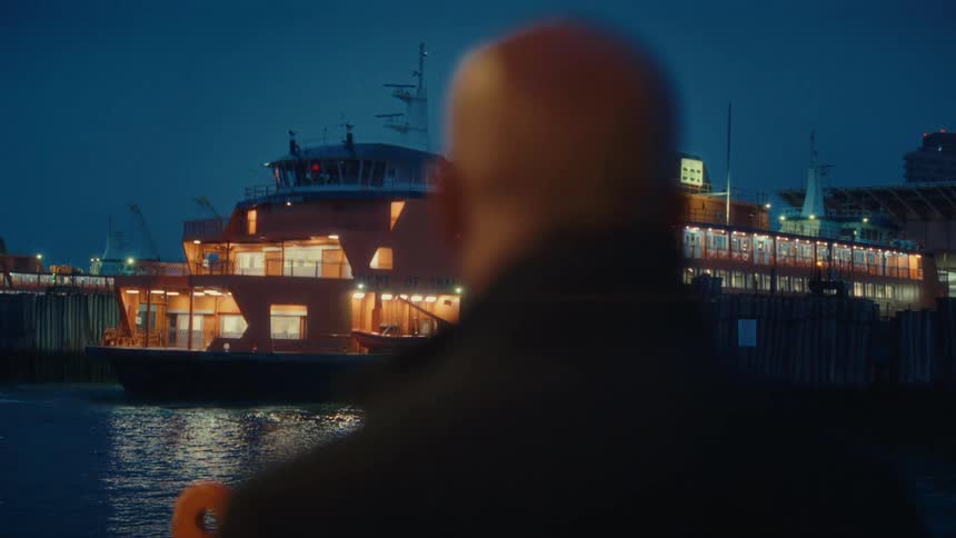 This nighttime video begins with a closeup shot of a bald man with a mustache looking thoughtfully toward the side with blurry orange bokeh lights visible in the backgroundthe man then turns to face the water revealing a large distinctive orange