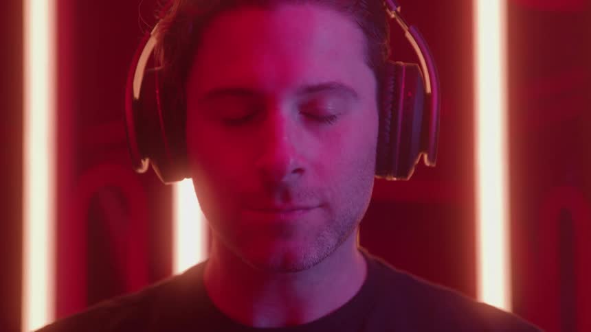 A man in a vibrant neon redlit environment first places a pair of overear headphones on his head and then closes his eyes with a slight smile