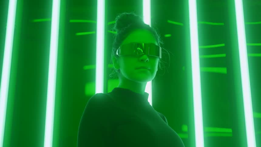 A woman with hair in a bun wears reflective futuristic visor glasses and a dark turtleneck looking forward against a background of bright vertical green neon lights