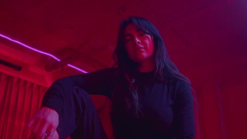 A stylish woman with long dark hair wearing all black and combat boots sits in a dark room illuminated by intense red and pink neon light