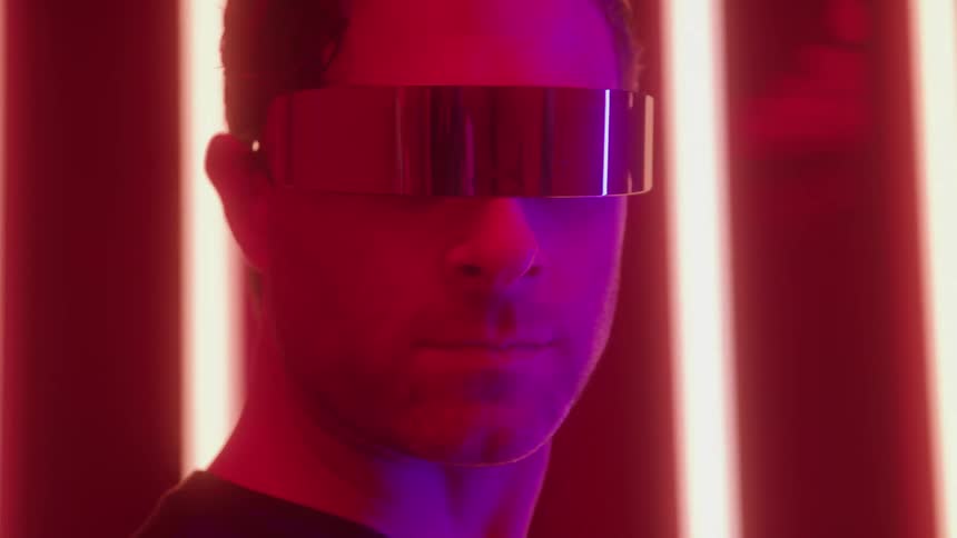 A man with a beard wearing mirrored futuristic visor glasses is illuminated by red and purple light against a background of vertical neon lights