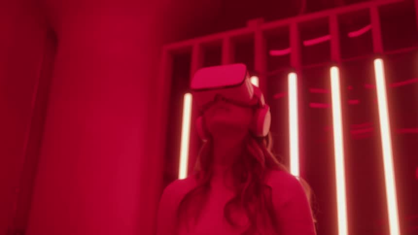A woman wearing a vr headset and headphones is illuminated by a strong reddishpink light in a dramatic setting behind her are vertical neon or fluorescent light tubes encased in a cagelike structure