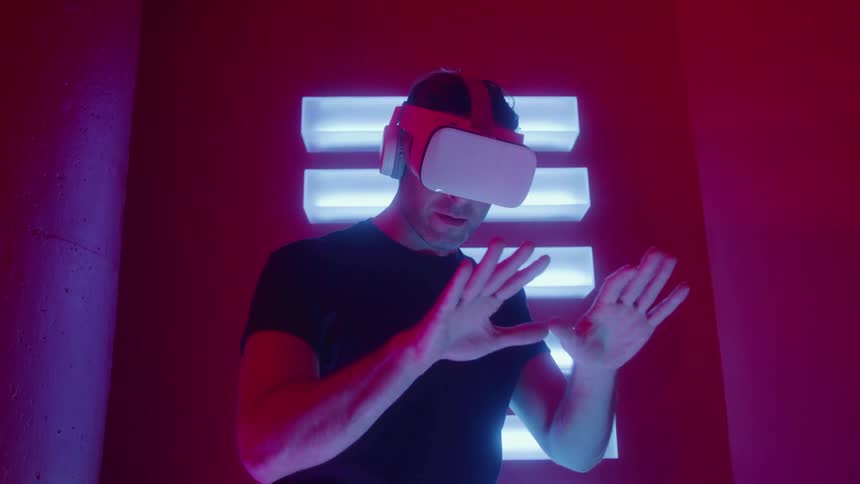 The video shows a man wearing a white and black virtual reality vr headset standing in an intensely lit room bathed in vibrant red and blue neon lighthe is interacting with the virtual environment by moving his hands in front of him suggesting he i