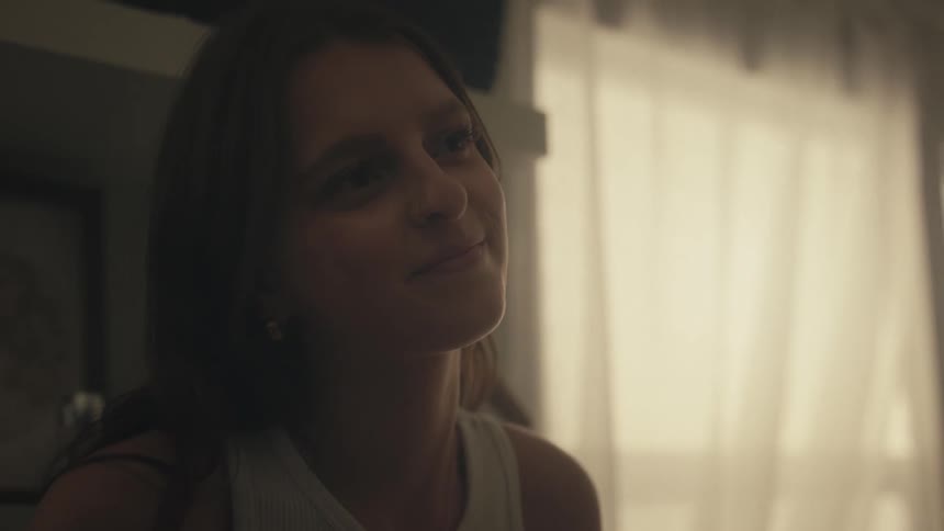 A close view of a young woman with long brown hair smiling and adjusting her hair in a dimly lit room with bright white curtains in the background