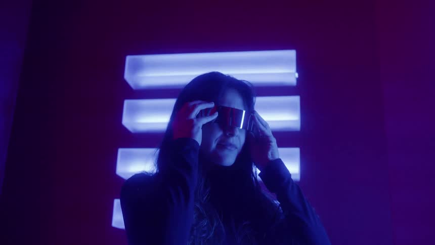 A woman with long dark hair puts on sleek futuristic glasses in a dark room illuminated by intense pink and blue neon lights that her hair back