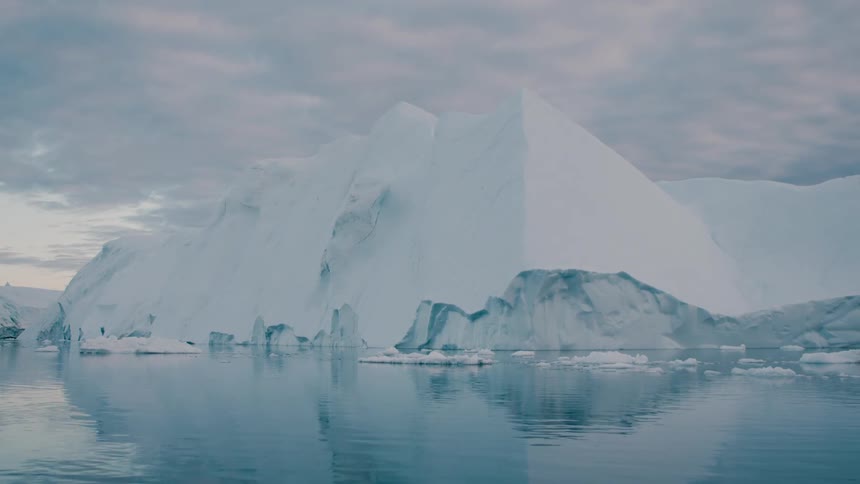 A massive white iceberg floats on calm blue water reflecting its angular shape against a pale gray and cloudy sky