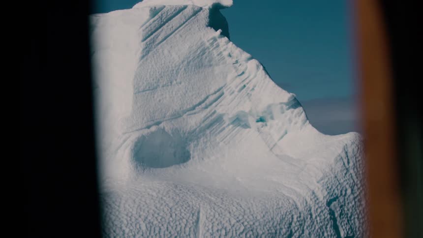 A massive snowcovered ice mountain and glaciers are visible against a bright blue sky in a body of water framed partially by a dark border in the foreground