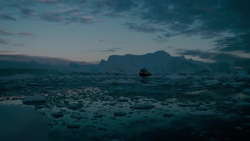 A small red and white vessel is visible far away in the dark ocean surrounded by chunks of ice and large icebergs under a dramatic cloudy twilight sky