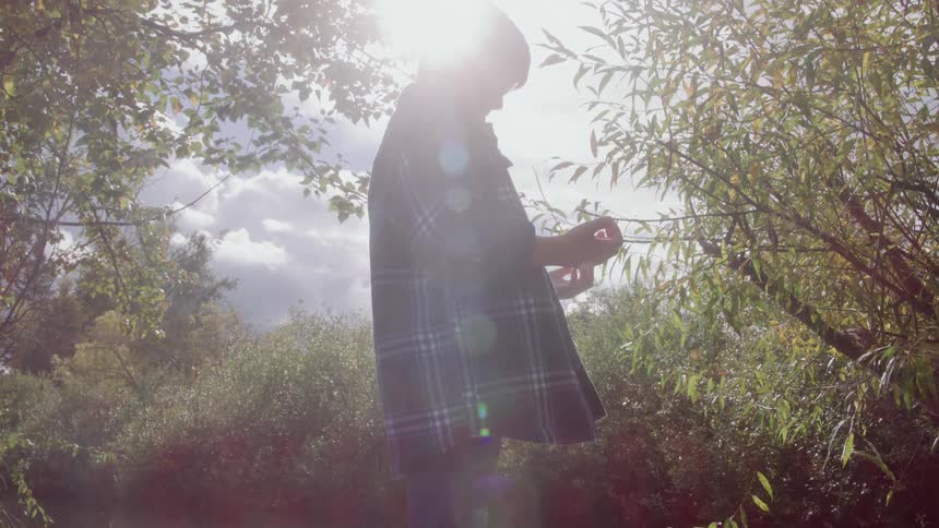 A boy wearing a plaid shirt is silhouetted against a bright sundrenched sky and green foliage performing an action with their hands in the natural outdoor light