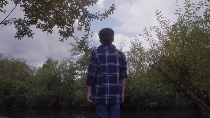 A young boy in a blue plaid shirt stands facing a calm stream surrounded by lush green and autumn foliage under a bright cloudy sky