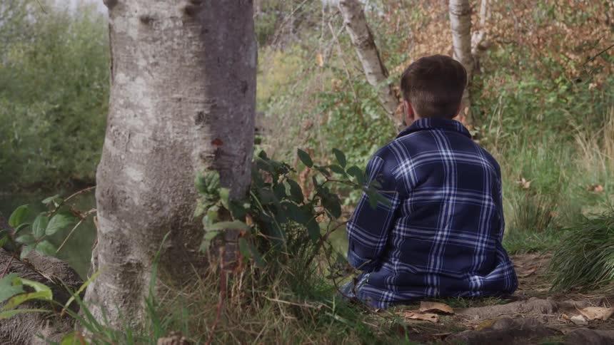 Boy sitting on the ground leaning against a tree in a wooded area
