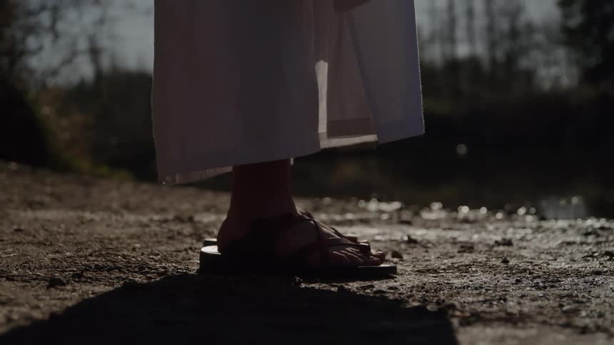 A low view focuses on a persons feet in brown sandals under a flowing white robe standing on dirt ground near water in soft morning light