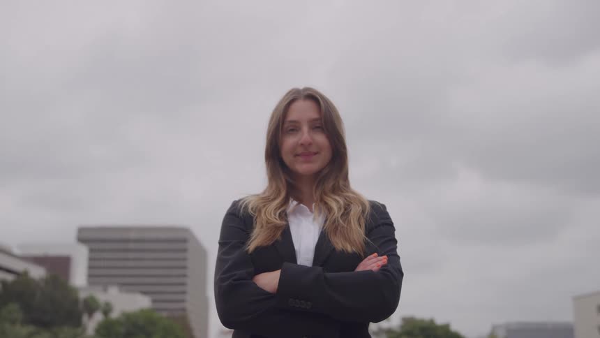 A young woman in a black business suit and white shirt stands outdoors in an urban area with a gray sky and buildings in the background turning to the camera with a confident smile and crossed arms