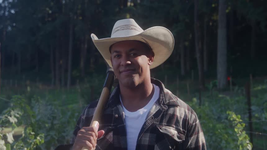 Young black gardener wearing a cowboy hat holding a tool on his shoulder and smiling next to a garden covered in plants with a forest on the background outdoors during day time