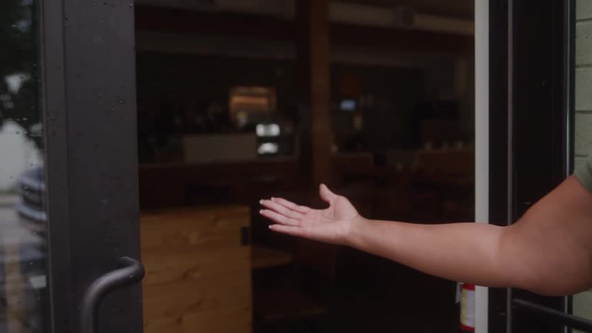 Young black woman opening the door to a restaurant with a smile and signing inside in an welcoming fashion outdoors during day time
