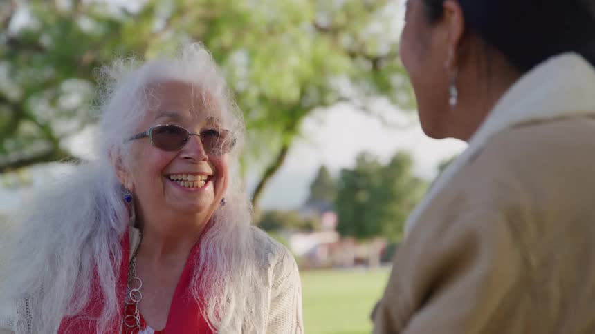 Elderly woman wearing sunglasses smiling and talking to a younger woman in a park outdoors