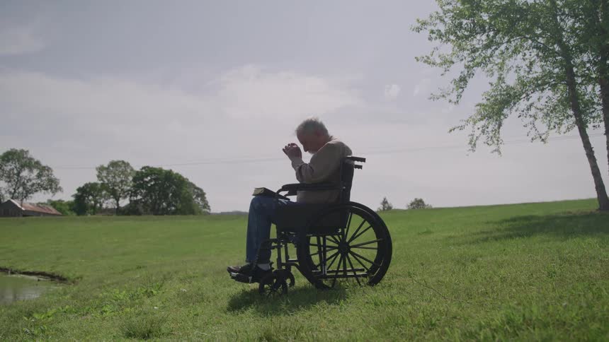 An older man in a wheelchair prays in a grassy outdoor area under a bright sky
