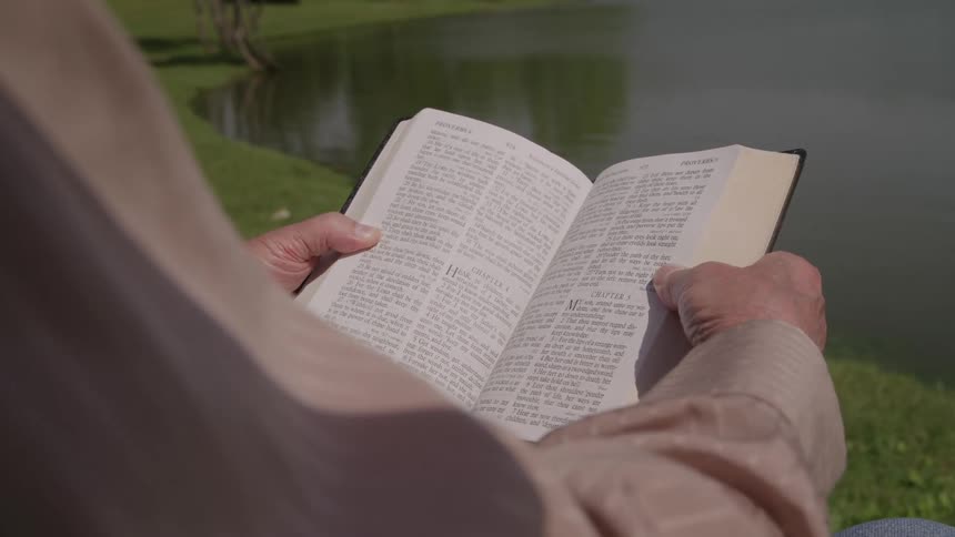 A close over the shoulder view shows an older person wearing a light brown shirt holding an open bible and reading aloud by the edge of a calm lake on a sunny day