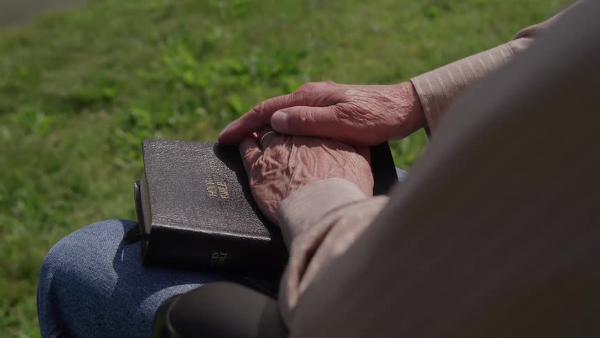 A closeup shot shows a persons hands resting on a black book labeled holy bible while sitting outdoors the person appears to be wearing blue jeans and a lightcolored longsleeve shirt with green grass visible in the background