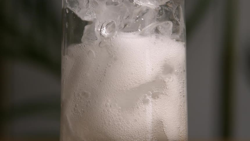 A close view of a clear glass filled with ice and a frothy carbonated beverage showing bubbles rapidly rising to the surface and a creamy white foam forming