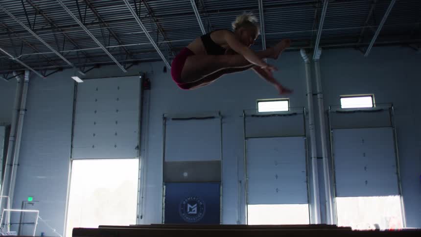 A female athlete is captured during a jump in a large brightly lit indoor gymnasium with high ceilings and large overhead doors