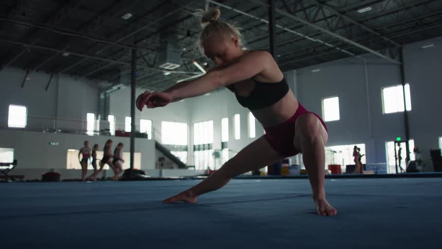 A female athlete with blonde hair tied in a bun is performing a deep side lunge or stretching exercise on a blue gymnasium floor she is wearing a black sports bra and maroon shorts and is leaning forward with her arms extended